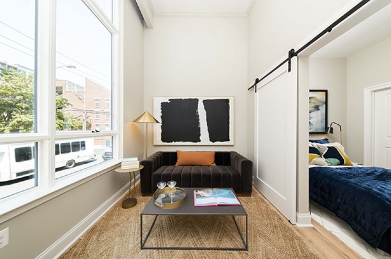 A furnished living room with large windows on the left wall and a large sliding door on the right wall. The sliding door is open to a furnished bedroom.