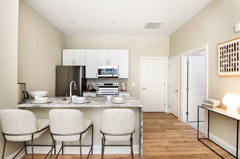 A kitchen aisle with 3 chairs on the living room side and a view of the cabinets, a refrigerator, a stove, a microwave on the opposite wall.  Doors to adjacent rooms are shown next to the kitchen area