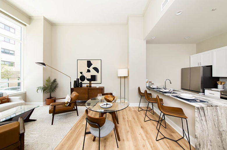 A furnished living room with a large window and a dining area on a kitchen aisle in an adjacent furnished kitchen