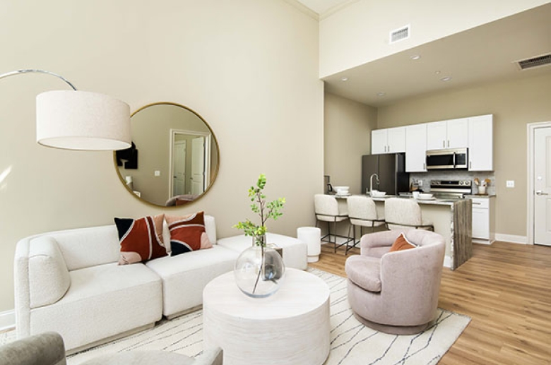 A furnished living room with an adjacent dining area on the kitchen aisle and a furnished kitchen in the background.