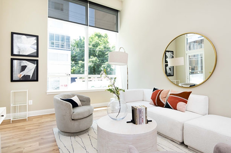 A bright furnished living room with a large window with sheer-weave blinds. To the right of the window there is a perpendicular wall with a round mirror reflecting the glass door to the patio/balcony on the opposite wall.