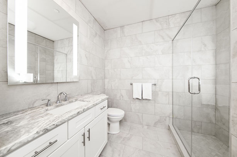 A tiled bathroom with a shower enclosed with a glass door. Across from the shower is a toilet and next to it a sink counter with drawers, and cabinets underneath and a large wall mirror above the sink.