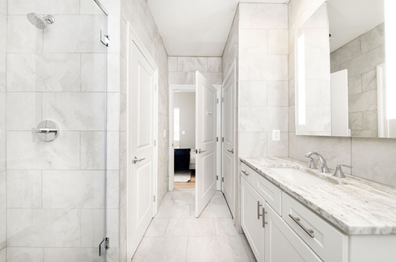 A view from inside of a bathroom.  On the left:  a shower head behind the glass door next to a closed door.  On the right:  a sink counter with cabinets and drawers underneath, with a large wall mirror above the sink.  In the center: a bathroom door opening into a bedroom. 