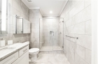 A bathroom view showing a toilet and a mirror cabinet above it, next to a sink with cabinets and a drawer, and a large mirror above the sink on the left and a tiled shower with glass door in the center