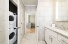 A bathroom with a sink with cabinets and drawers and large wall mirror on the right side and stackable washer and dryer in a closet on the left side, with the closet door open.  An open bathroom door in the center shows a view of a kitchen aisle in the background. 