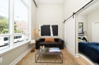 A furnished living room with large windows on the left wall and a large sliding door on the right wall. The sliding door is open to a furnished bedroom.