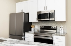 Kitchen cabinets, a large refrigerator, a microwave, and a stove with tiles on the wall behind it, all placed along one wall of the kitchen, across from the kitchen aisle.