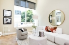 A bright furnished living room with a large window with sheer-weave blinds. To the right of the window there is a perpendicular wall with a round mirror reflecting the glass door to the patio/balcony on the opposite wall.